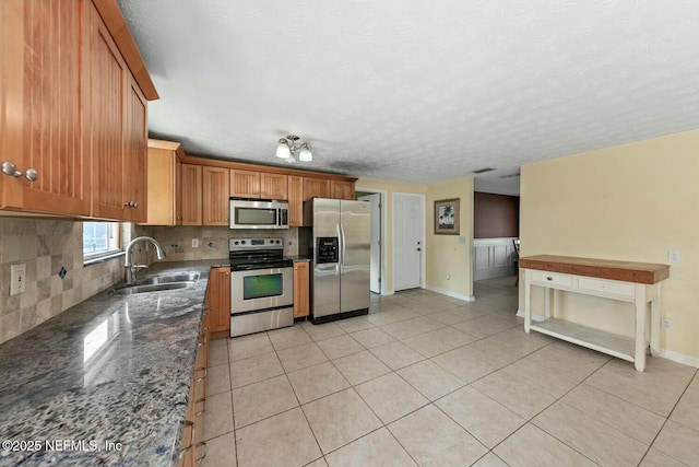 kitchen with appliances with stainless steel finishes, a sink, dark stone counters, light tile patterned floors, and backsplash