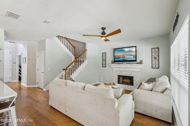 living area with a brick fireplace, a ceiling fan, wood finished floors, visible vents, and stairway