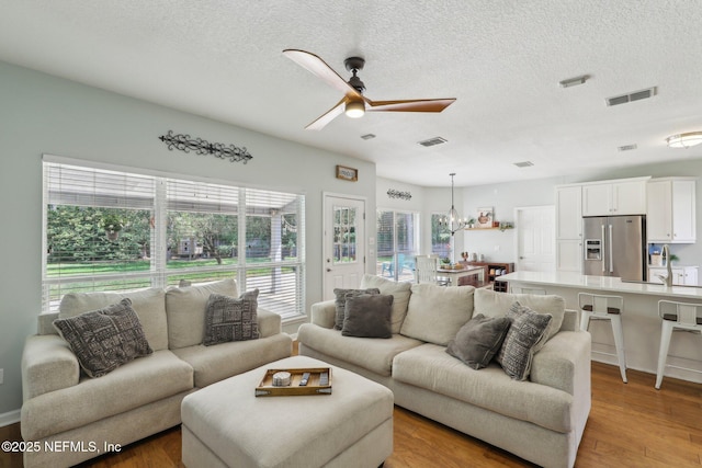 living area with ceiling fan with notable chandelier, light wood finished floors, a textured ceiling, and visible vents