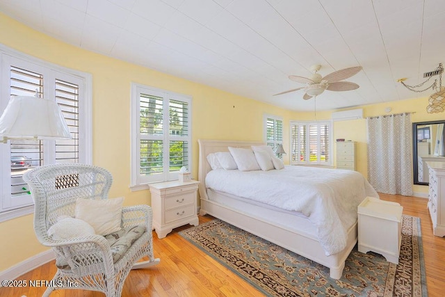 bedroom featuring a wall unit AC, multiple windows, light wood-style flooring, and ceiling fan