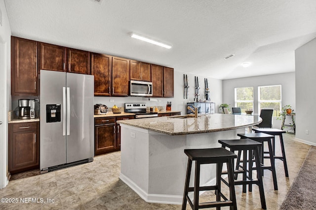 kitchen featuring light stone counters, a sink, stainless steel appliances, a kitchen bar, and a center island with sink