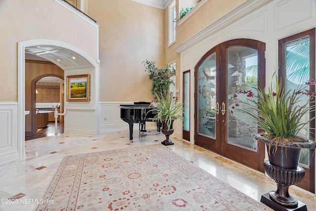 foyer featuring a decorative wall, arched walkways, french doors, light marble finish floors, and crown molding