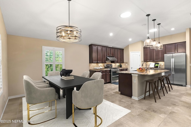 kitchen with dark brown cabinets, under cabinet range hood, a chandelier, tasteful backsplash, and stainless steel appliances