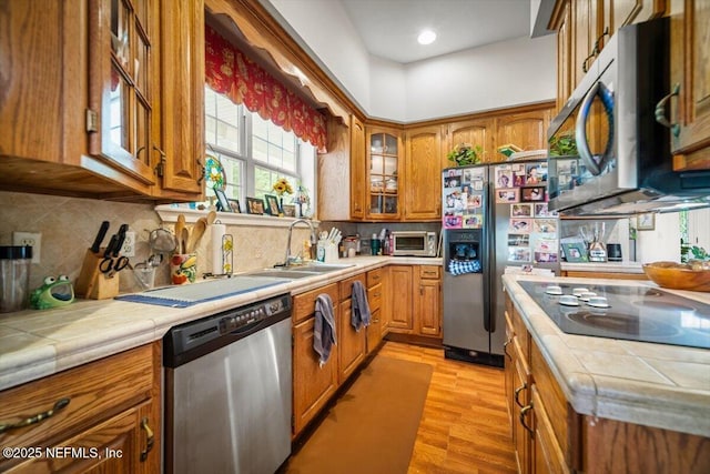 kitchen with glass insert cabinets, a sink, tile counters, stainless steel appliances, and brown cabinetry