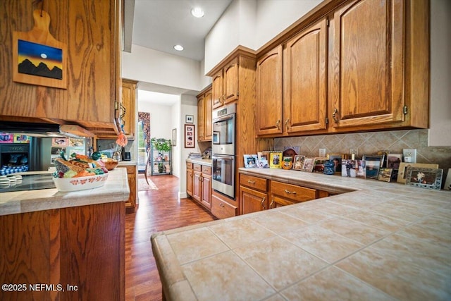 kitchen featuring brown cabinets, decorative backsplash, tile countertops, double oven, and wood finished floors