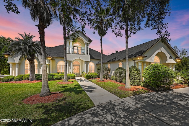 traditional-style house featuring stucco siding, a yard, a balcony, and a shingled roof