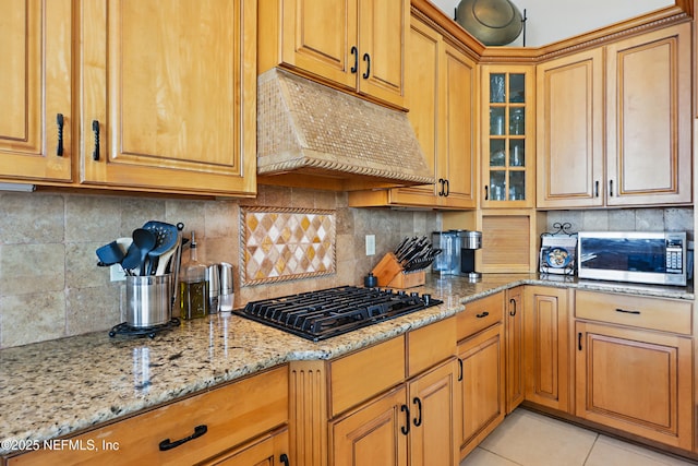kitchen featuring stainless steel microwave, glass insert cabinets, under cabinet range hood, light stone countertops, and black gas stovetop