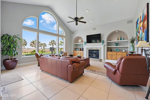 living room with light tile patterned floors, a glass covered fireplace, a ceiling fan, high vaulted ceiling, and built in shelves