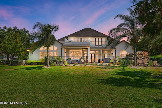 back of house at dusk featuring a lawn, fence, a balcony, and stucco siding