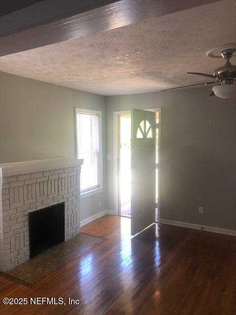 unfurnished living room with a brick fireplace, wood finished floors, a ceiling fan, baseboards, and a textured ceiling