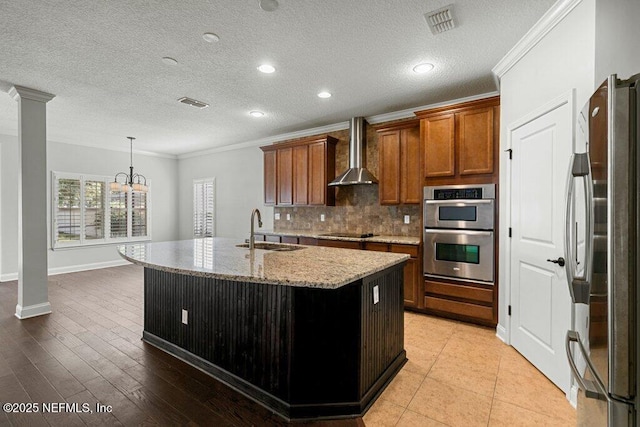 kitchen with stainless steel appliances, wall chimney exhaust hood, crown molding, a sink, and an island with sink