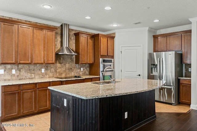 kitchen with stainless steel appliances, wall chimney range hood, a sink, light stone countertops, and an island with sink