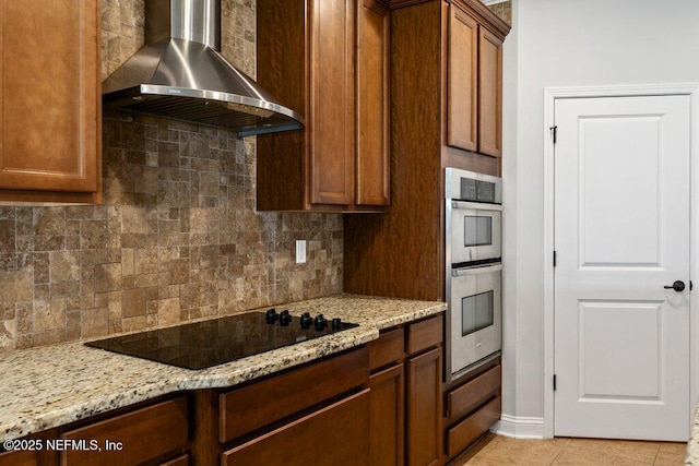 kitchen with wall chimney range hood, black electric stovetop, tasteful backsplash, light stone countertops, and double oven