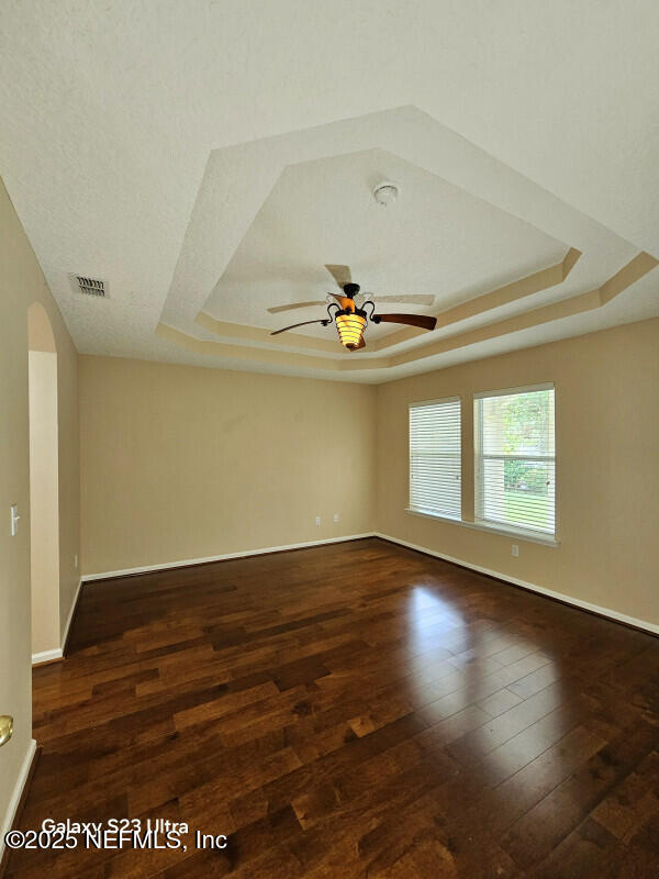 empty room with a ceiling fan, wood finished floors, a tray ceiling, baseboards, and visible vents