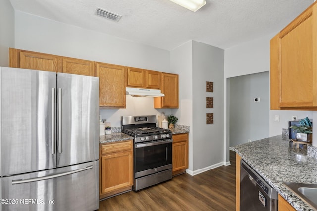 kitchen with stainless steel appliances, under cabinet range hood, dark wood-type flooring, stone counters, and visible vents