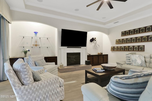 living room featuring ceiling fan, light wood-style floors, a tray ceiling, arched walkways, and a glass covered fireplace