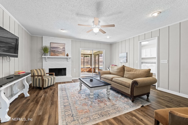 living room with crown molding, a decorative wall, a brick fireplace, a ceiling fan, and dark wood-type flooring