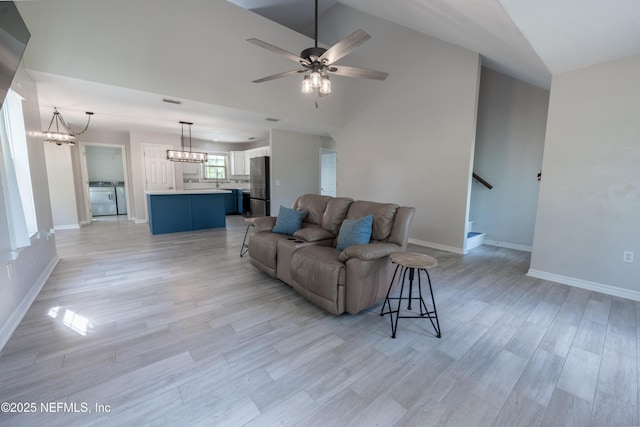 living room featuring lofted ceiling, washing machine and dryer, light wood-style floors, baseboards, and ceiling fan with notable chandelier