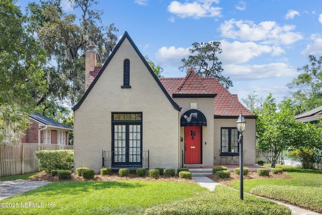 tudor-style house with fence, stucco siding, a chimney, and a front yard
