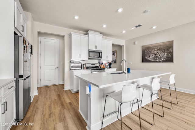 kitchen featuring appliances with stainless steel finishes, a sink, a center island with sink, light wood finished floors, and a breakfast bar