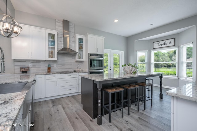 kitchen with decorative backsplash, a kitchen breakfast bar, wall chimney range hood, double oven, and white cabinets