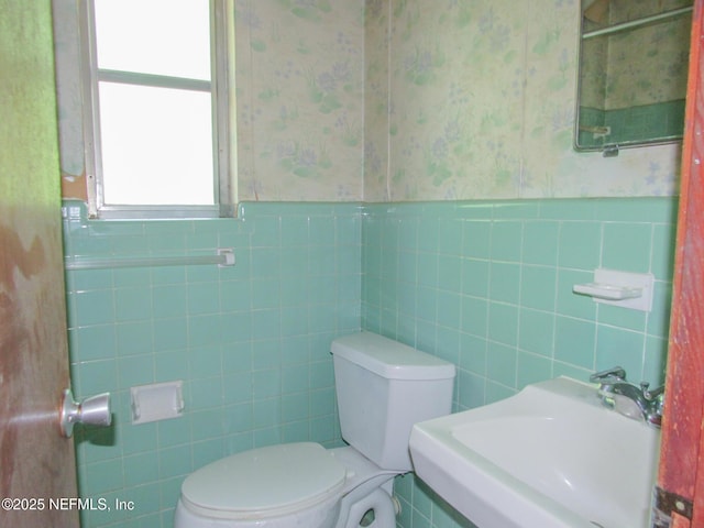 bathroom with toilet, tile walls, wallpapered walls, a wainscoted wall, and a sink