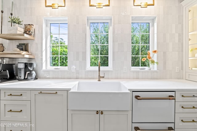 kitchen with open shelves, white dishwasher, light countertops, a sink, and backsplash