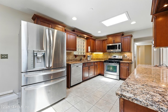kitchen with light tile patterned flooring, light stone countertops, appliances with stainless steel finishes, brown cabinetry, and backsplash