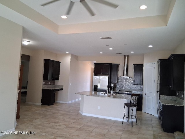 kitchen with wall chimney range hood, backsplash, appliances with stainless steel finishes, and a kitchen island with sink