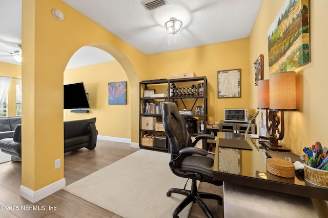 home office featuring arched walkways, a textured ceiling, baseboards, wood finished floors, and visible vents
