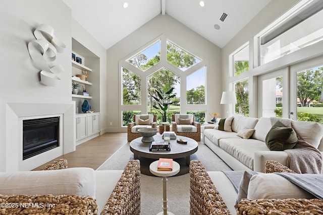 living area with high vaulted ceiling, a glass covered fireplace, plenty of natural light, and light wood-style floors