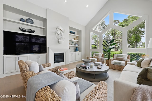 living area with a glass covered fireplace, high vaulted ceiling, light wood finished floors, and built in shelves