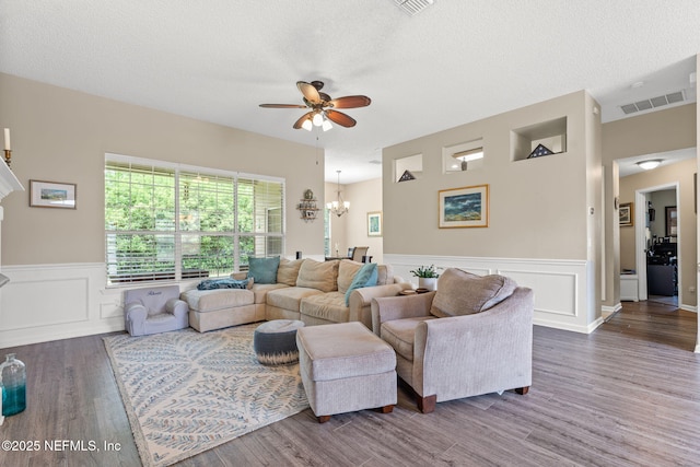 living area with visible vents, wood finished floors, a decorative wall, a wainscoted wall, and ceiling fan with notable chandelier