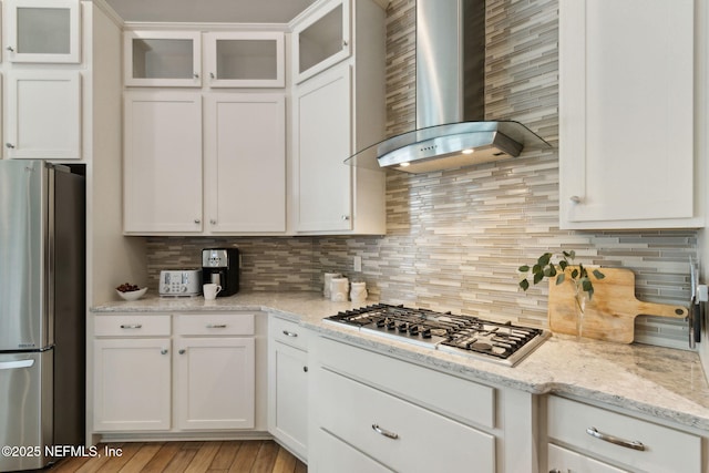 kitchen featuring wall chimney range hood, stainless steel appliances, white cabinetry, backsplash, and glass insert cabinets