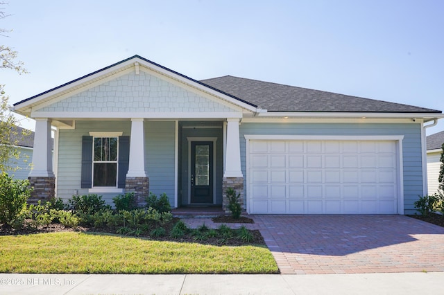 craftsman-style house featuring covered porch, a front yard, decorative driveway, an attached garage, and a shingled roof