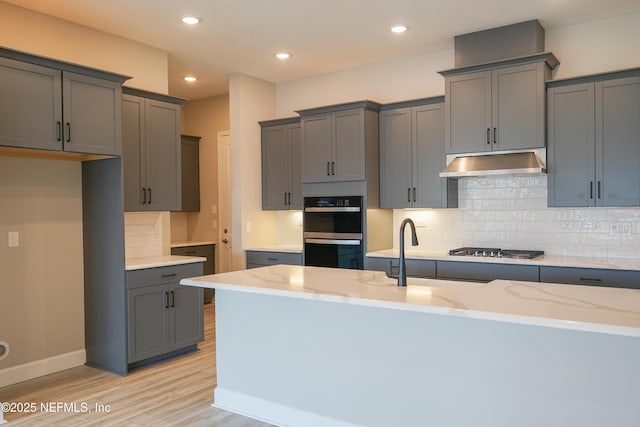 kitchen with under cabinet range hood, tasteful backsplash, light stone counters, light wood finished floors, and double wall oven