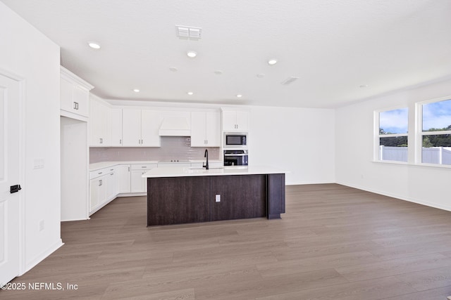 kitchen with oven, light wood-style floors, a center island with sink, light countertops, and built in microwave
