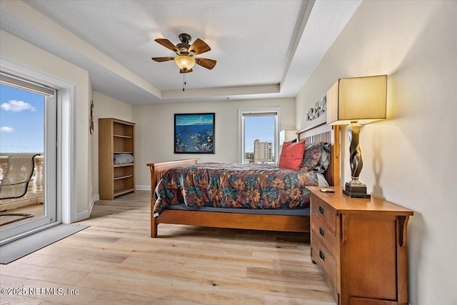 bedroom with access to outside, a tray ceiling, light wood-style floors, baseboards, and a ceiling fan