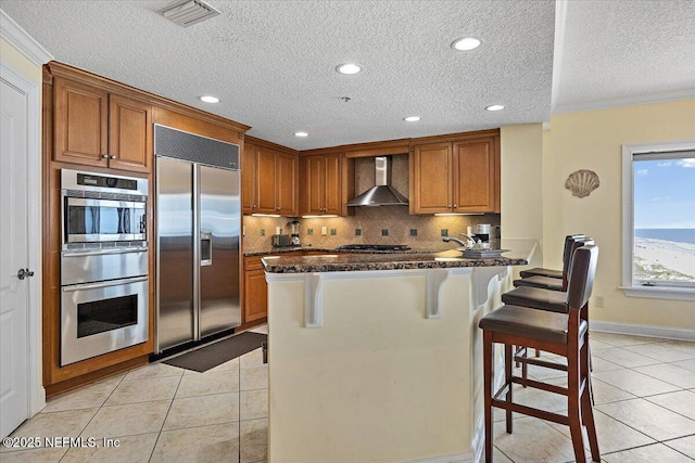 kitchen featuring stainless steel built in refrigerator, wall chimney exhaust hood, light tile patterned floors, a breakfast bar, and a warming drawer