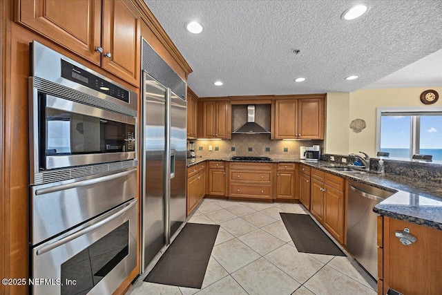 kitchen with wall chimney exhaust hood, built in appliances, a sink, brown cabinetry, and a textured ceiling