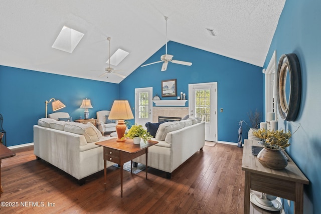 living room featuring a ceiling fan, dark wood-style flooring, a tiled fireplace, baseboards, and lofted ceiling with skylight