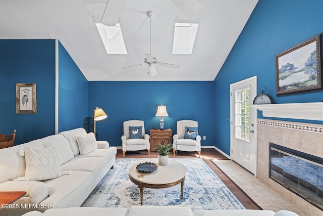 living room featuring vaulted ceiling with skylight, a tiled fireplace, a ceiling fan, baseboards, and wood finished floors