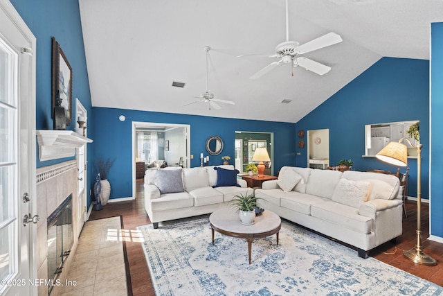 living room with visible vents, baseboards, high vaulted ceiling, a tiled fireplace, and a ceiling fan