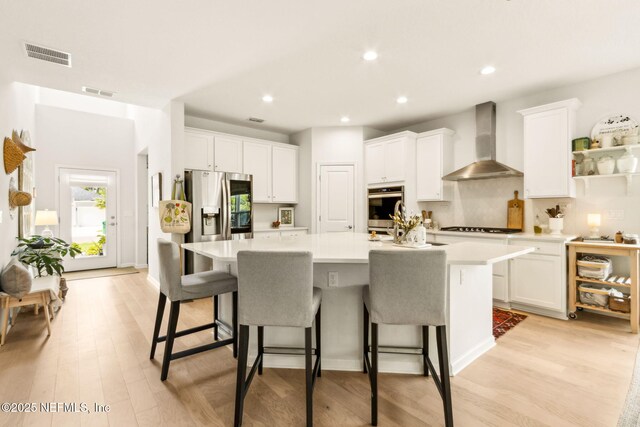 kitchen featuring wall chimney range hood, white cabinets, visible vents, and an island with sink