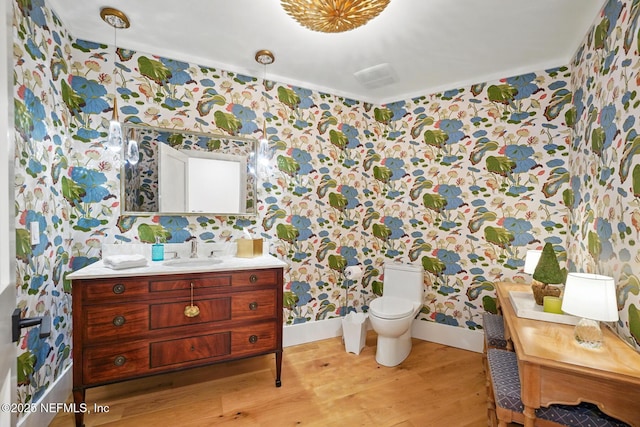 bathroom featuring vanity, wood finished floors, toilet, baseboards, and wallpapered walls