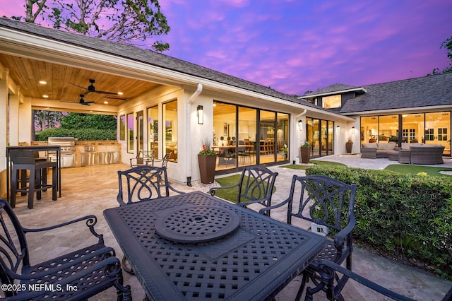 patio terrace at dusk featuring a ceiling fan, outdoor dining area, an outdoor kitchen, outdoor lounge area, and a grill