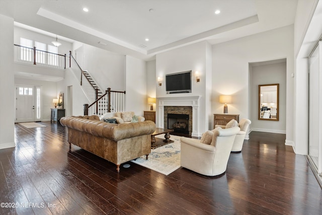 living area with dark wood-style flooring, a tray ceiling, a stone fireplace, and baseboards