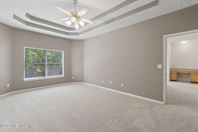 empty room featuring a raised ceiling, ceiling fan, baseboards, and light colored carpet
