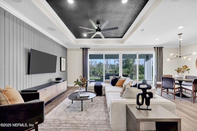 living room featuring ornamental molding, baseboards, light wood-type flooring, a raised ceiling, and ceiling fan with notable chandelier