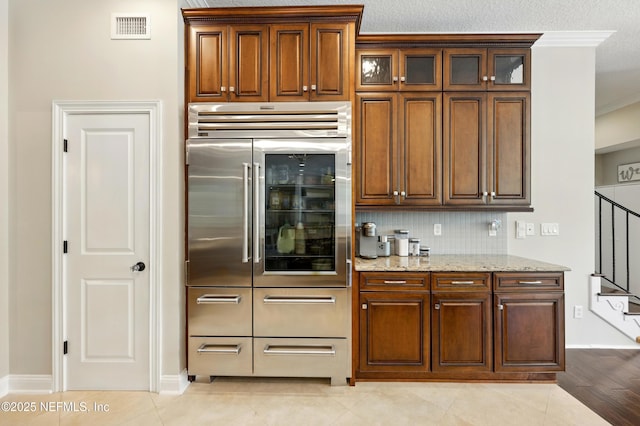 kitchen featuring stainless steel built in fridge, light stone counters, decorative backsplash, ornamental molding, and glass insert cabinets